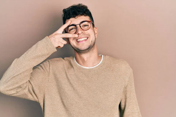 Gündelik kıyafetler giyen ve gözlük takan genç İspanyol adam barış sembolü yapıyor. Yüzünde parmaklar, gülümseyerek zaferi gösteriyor. 