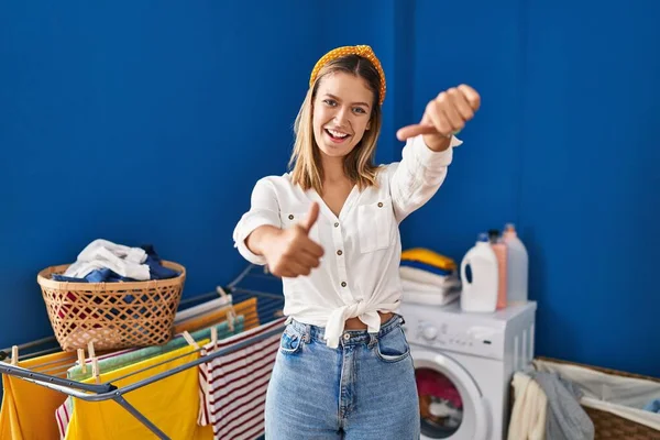 Young blonde woman at laundry room approving doing positive gesture with hand, thumbs up smiling and happy for success. winner gesture.