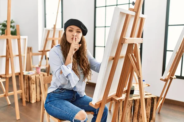 若いヒスパニックアーティストの女性は アートスタジオでキャンバスに絵を描く唇に指で静かにするように求めています 沈黙と秘密の概念 — ストック写真