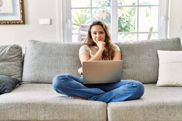 Kanepede oturan genç ve güzel esmer bir kadın bilgisayarlı dizüstü bilgisayar kullanarak kameraya bakıyor kollarını kavuşturup gülümseyerek ve çenesini kaldırarak bakıyor. Olumlu düşünmek. 