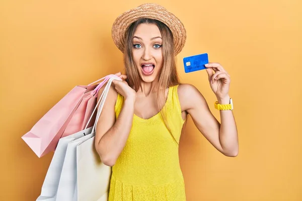 Young blonde girl holding shopping bags and credit card celebrating crazy and amazed for success with open eyes screaming excited.