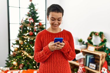 Genç İspanyol kadın akıllı telefon kullanıyor. Noel ağacının yanında duruyor.