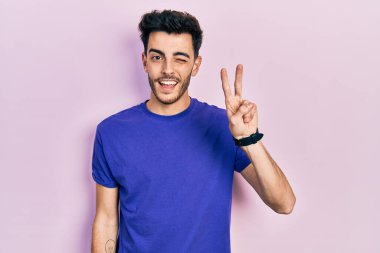 Gündelik tişört giyen, gülümseyen ve kameraya göz kırpan genç İspanyol adam parmaklarıyla zafer işareti yapıyor. İki numara.. 