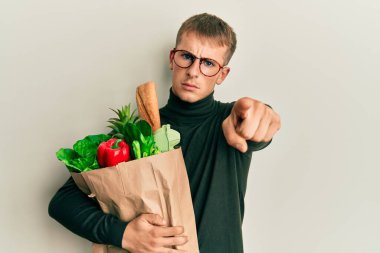 Elinde alışveriş poşeti olan genç beyaz bir adam kamerayı işaret ediyor ve kendine güveni tam bir jest yapıyorsun. 