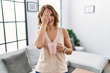 Orta yaşlı bir kadın evde kahve içiyor. Bir gözünü eliyle kapatıyor. Yüzünde kendine güvenen bir gülümseme ve sürpriz bir duygu var.. 