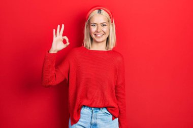 Gündelik kırmızı kazak giyen güzel sarışın kadın gülümsüyor ve el ve parmaklarıyla 