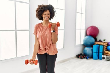 Genç Afrikalı Amerikalı kadın gülümsüyor spor merkezinde halterleri kullanarak kendine güven eğitimi alıyor