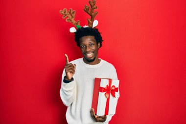 Young african american man wearing deer christmas hat holding gift pointing finger up with successful idea. exited and happy. number one. 