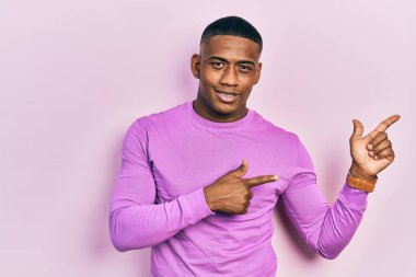 Genç siyah bir adam pembe bir süveter giyiyor gülümsüyor ve kameraya bakıyor iki eliyle ve parmaklarıyla kenarı işaret ediyor.. 