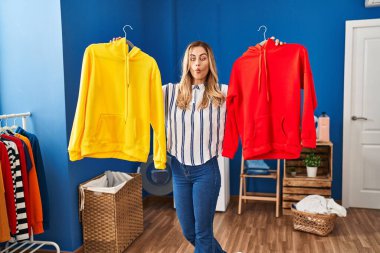 Young blonde woman holding clean laundry on hangers making fish face with mouth and squinting eyes, crazy and comical. 