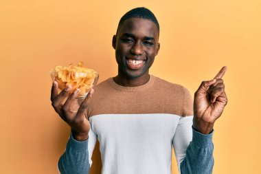 Genç Afro-Amerikan adam patates cipsiyle kaseyi tutarken mutlu bir şekilde gülümsüyor. 