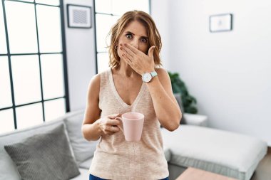 Evde kahve içen orta yaşlı bir kadın yanlışlıkla elleriyle ağzını kapatırken şok oldu. gizli kavram. 