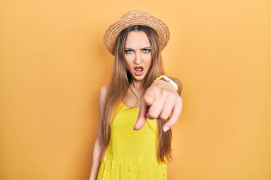 Young blonde girl wearing summer hat pointing displeased and frustrated to the camera, angry and furious with you 