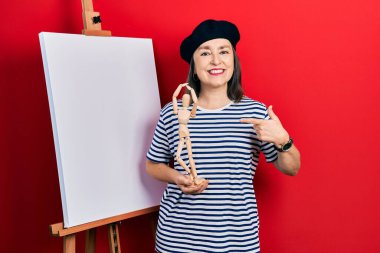 Orta yaşlı İspanyol kadın, resim sehpasının yanında durup elinde manikinle gülümsüyor ve mutlu bir şekilde elini ve parmağını işaret ediyor. 