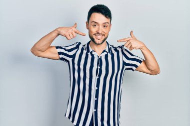 Günlük kıyafetler giyen, neşeli gülümseyen, parmaklarıyla ve ağzıyla işaret eden İspanyol genç bir adam. diş sağlığı kavramı. 