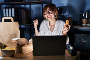 Genç ve güzel bir kadın bilgisayar laptopu kullanıyor ve yemek yiyor çok mutlu ve heyecanlı bir şekilde kollarını kaldırıp, gülümseyerek ve başarı için çığlık atarak zafer jesti yapıyor. kutlama konsepti. 
