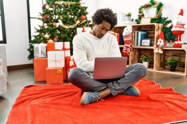 Genç Afro-Amerikan adam dizüstü bilgisayar kullanıyor. Noel ağacının yanında oturuyor. Kuşkucu ve gergin. negatif kişi. 