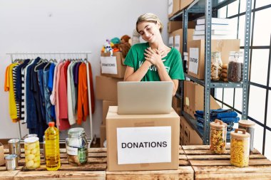 Young blonde woman wearing volunteer t shirt at donations stand smiling with hands on chest with closed eyes and grateful gesture on face. health concept. 