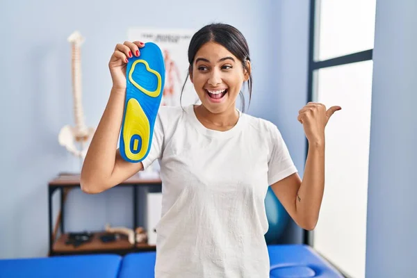 Young hispanic woman holding shoe insole at physiotherapy clinic pointing thumb up to the side smiling happy with open mouth