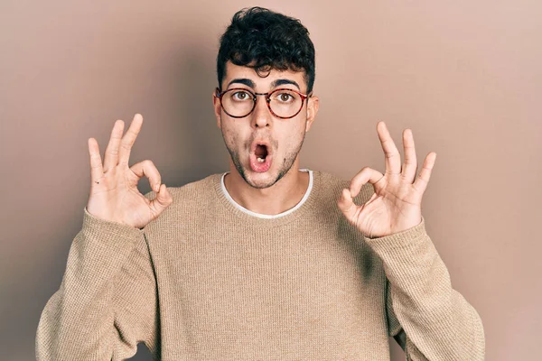 Gündelik kıyafetler ve gözlük takan genç İspanyol adam şaşırmış ve şok olmuş bir şekilde parmaklarıyla onay sembolü yapıyor. çılgın ifade 