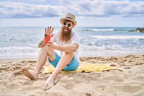 Genç kızıl saçlı adam sahilde havlunun üzerinde otururken akıllı telefonun yanında selfie çekiyor..