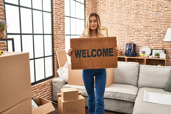 Young Blonde Woman Holding Welcome Doormat New Home Relaxed Serious — Stock fotografie
