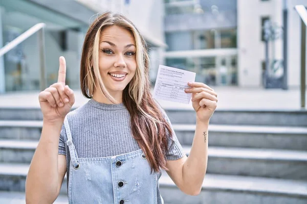 Young caucasian woman holding covid record card surprised with an idea or question pointing finger with happy face, number one