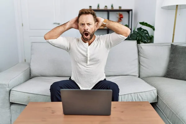 Middle age man using laptop at home crazy and scared with hands on head, afraid and surprised of shock with open mouth