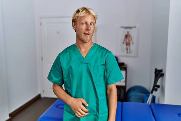 Jonge Blonde Man Draagt Fysiotherapeut Uniform Staan Kliniek Knipogen Naar — Stockfoto