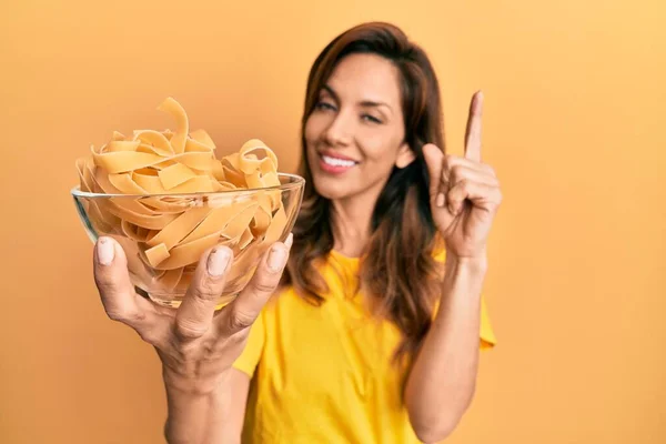 年轻的拉丁女人拿着碗和未煮熟的面食 开心地微笑着 手指头指向旁边 — 图库照片