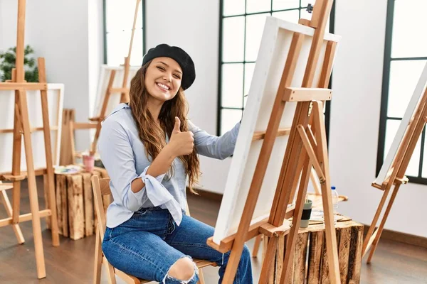 若いヒスパニック系のアーティストの女性は アートスタジオで手で幸せな親指をジェスチャーを行う上でキャンバスに絵を描く 成功を示すカメラを見ると — ストック写真