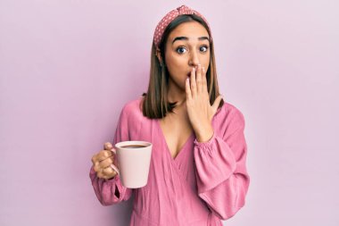 Genç esmer kız bir fincan kahve içiyor. Ağzını eliyle kapatıyor. Şok geçiriyor ve hata yapmaktan korkuyor. şaşırmış ifade 