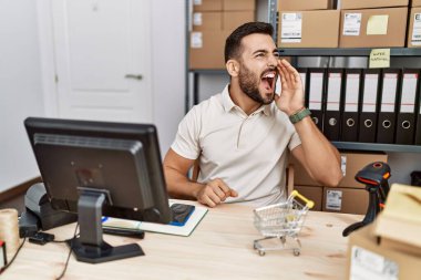 Handsome hispanic man working at small business commerce shouting and screaming loud to side with hand on mouth. communication concept. 