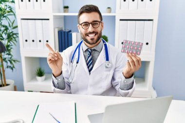 Yakışıklı, doktor üniforması giyen, elinde reçeteli haplarla gülümseyen, mutlu, eli ve parmağı yanda olan İspanyol bir adam. 