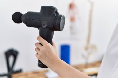 Klinikte perküsyon tabancası kullanan fizyoterapist üniforması giyen beyaz bir kadın.