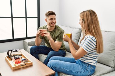 Genç İspanyol çift mutlu bir şekilde gülümsüyor. Evdeki kanepede oturup kahvaltı yapıyorlar..