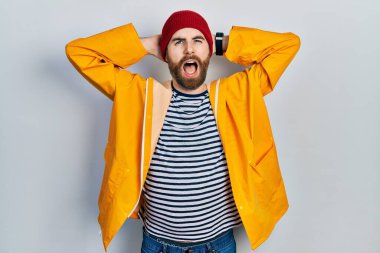 Caucasian man with beard wearing yellow raincoat crazy and scared with hands on head, afraid and surprised of shock with open mouth 