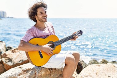 Genç İspanyol adam klasik gitar çalıyor plajda rock 'ın üzerinde oturuyor..