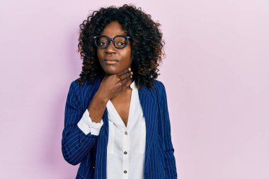 Genç Afrikalı Amerikalı kadın iş kıyafetleri ve gözlük takıyor. Ağrılı boynuna dokunuyor. Grip, mantar ve enfeksiyon yüzünden boğazı ağrıyor. 