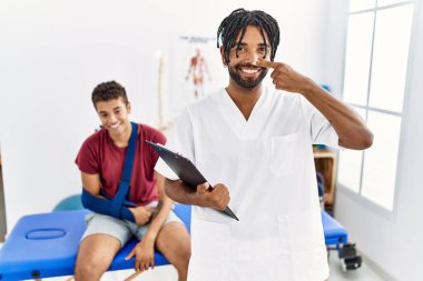 Kırık kollu bir adamla birlikte acı dindirme kliniğinde çalışan genç İspanyol bir adam. Parmakla yüzünü ve burnunu işaret ediyor. Neşeli bir şekilde gülümsüyor. güzellik konsepti 