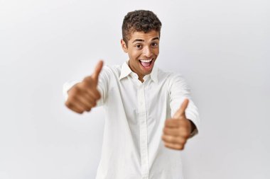 Genç, yakışıklı İspanyol bir adam izole edilmiş arka planda duruyor. Elle olumlu jest yapıyor, gülümsüyor ve başarı için mutlu. kazanan jest. 