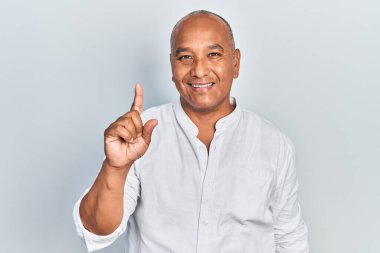 Orta yaşlı, gündelik kıyafetler giyen Latin bir numaralı parmağını gösterip işaret ederken kendinden emin ve mutlu bir şekilde gülümsüyor.. 