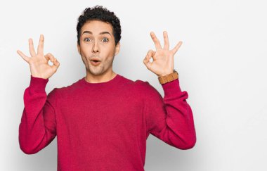 Gündelik kıyafetler giyen genç yakışıklı adam şaşırmış ve şok olmuş görünüyor. Tamam sembolünü parmaklarıyla yapıyor. çılgın ifade 