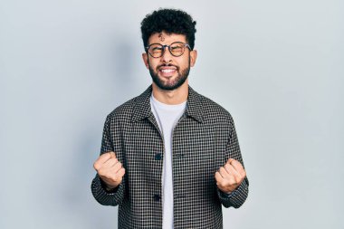 Sakallı genç Arap adam çok mutlu ve heyecanlı kolları havada, gülümseyen ve başarı için çığlık atan kazanan bir jest yapıyor. kutlama konsepti. 