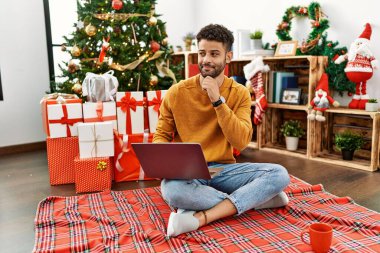 Arab young man using laptop sitting by christmas tree with hand on chin thinking about question, pensive expression. smiling and thoughtful face. doubt concept. 