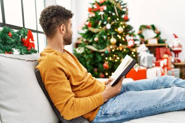 Genç Arap adam kitap okuyor. Evdeki Noel ağacının yanında, kanepede oturuyor..