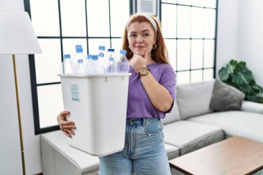 Geri dönüşüm sepetini elinde plastik şişelerle tutan genç kızıl saçlı kadın kameraya kollarını kavuşturmuş ve çenesini kaldırmış gülümsüyor. Olumlu düşünmek. 