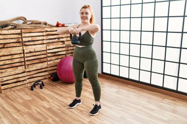 Genç kızıl saçlı kadın gülümsüyor spor merkezinde kettlebell ile antrenman yapıyor.