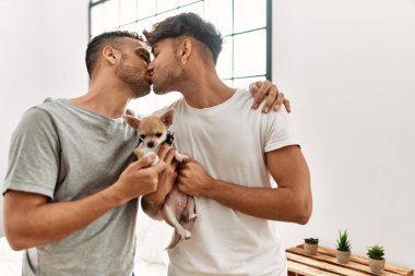 İki İspanyol erkek öpüşüyor ve sarılıyorlar. Yatak odasında chihuahua ile duruyorlar.