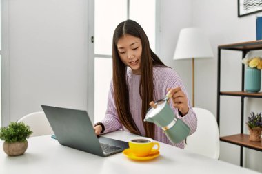 Genç Çinli kız dizüstü bilgisayar kullanıyor ve kahve içiyor. Evdeki masada oturuyor..